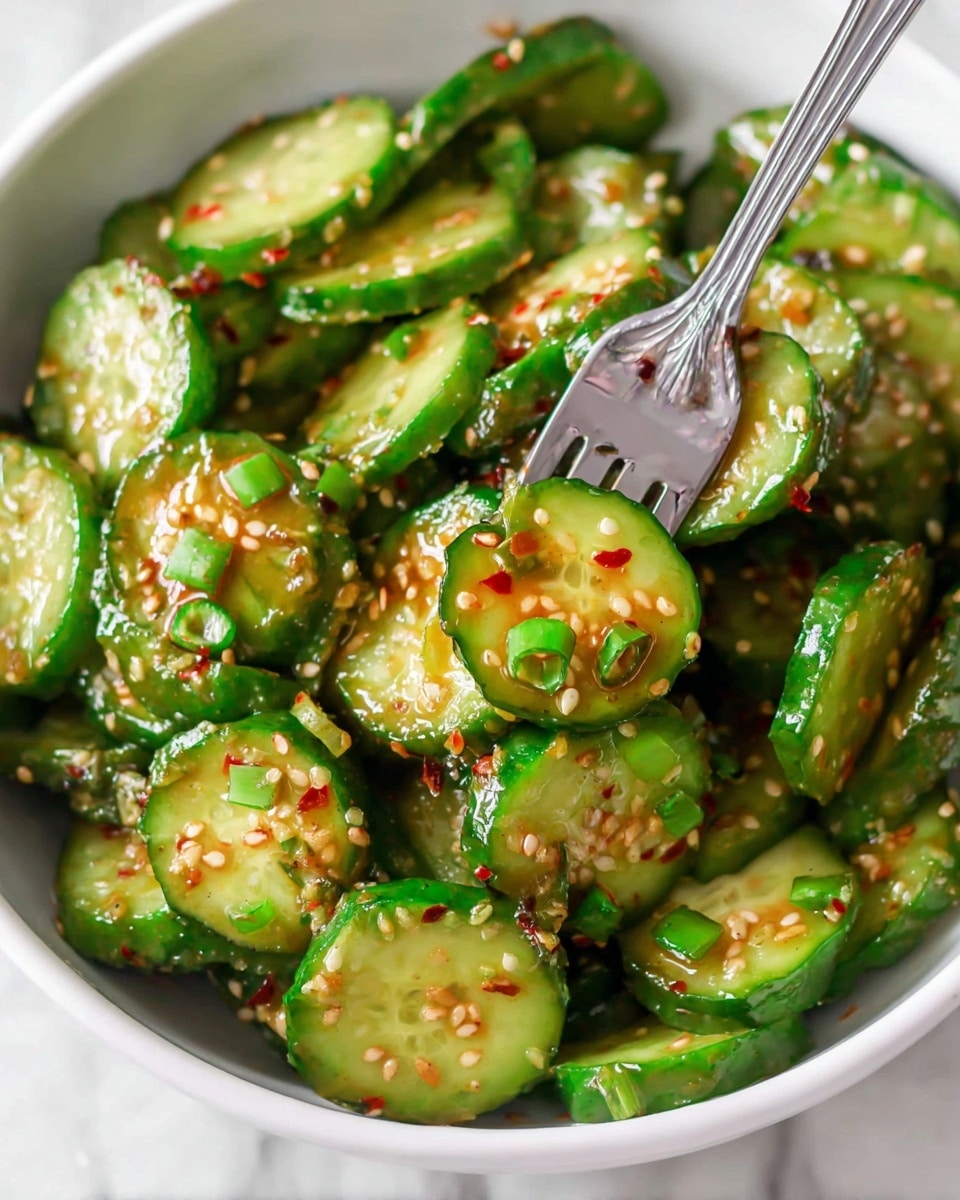 A bowl filled with many cucumber slices showing a bright green outer skin and light green inside with seeds, each slice coated with small specks of sesame seeds and red chili flakes. Scattered thin green onion pieces add different shades of green on top and throughout the cucumbers. A silver fork is seen among the cucumber slices, lifting a slice with a bit of sauce dripping from it. The bowl holding the cucumbers is white, and the background is a white marbled texture. photo taken with an iphone --ar 4:5 --v 7