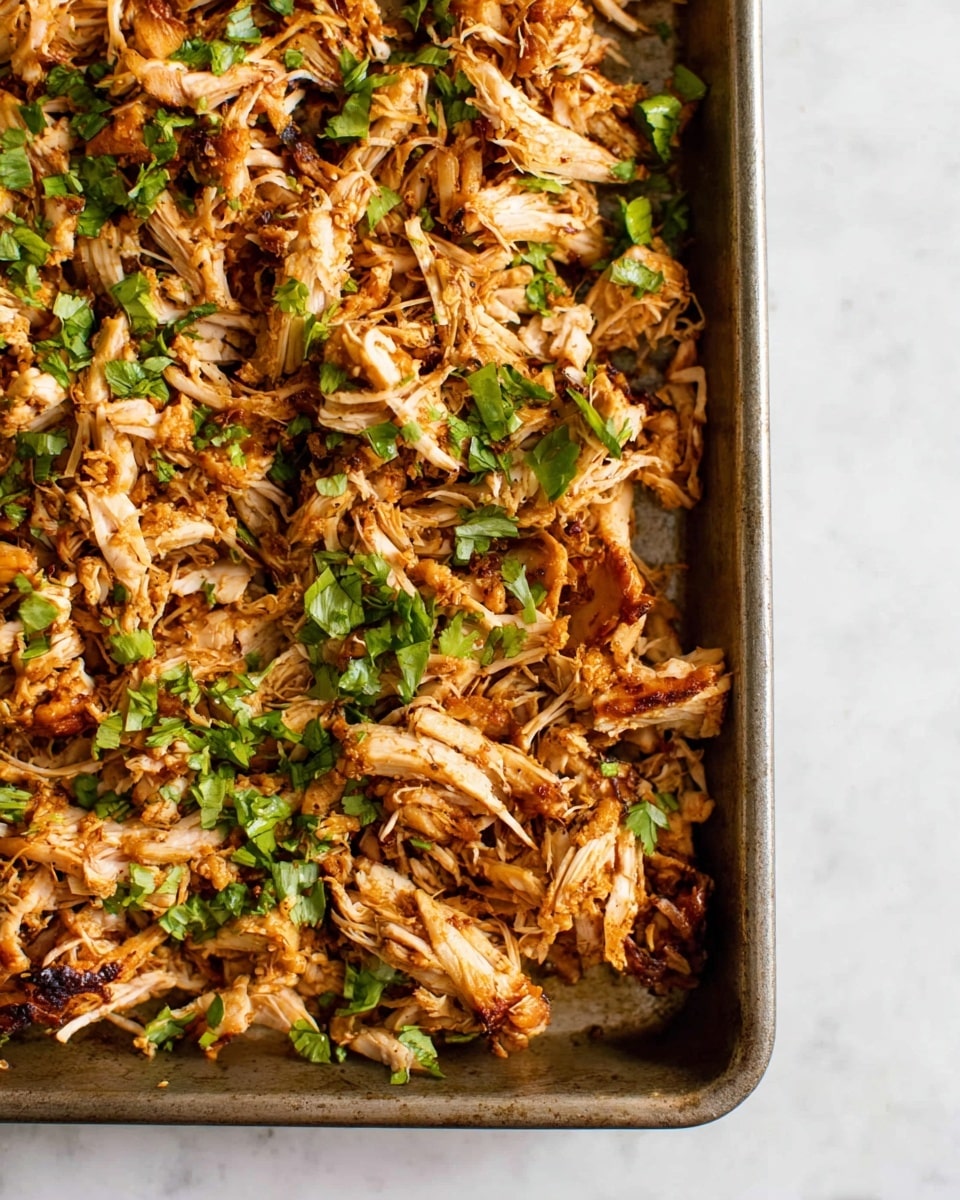 Close-up view of a baking tray filled with shredded cooked chicken that has a light brown and slightly charred color, showing toasted edges and some crispy parts. The top layer is scattered with fresh chopped green herbs, giving a bright contrast to the warm tones of the chicken. The tray is placed on a white marbled surface, and the texture of the chicken looks tender with some visible fibers and slight moisture. photo taken with an iphone --ar 4:5 --v 7