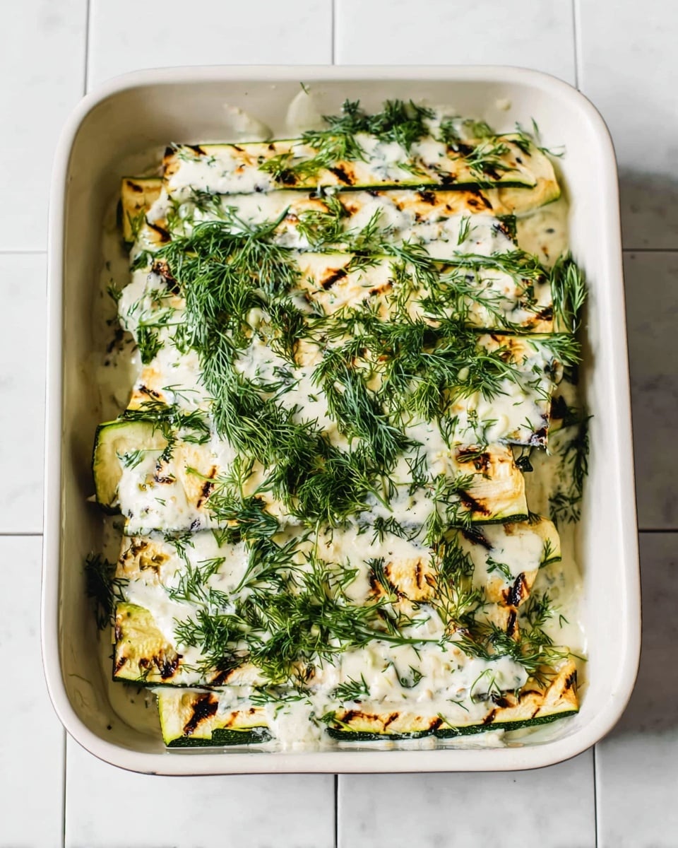The image shows a white rectangular baking dish filled with three stacked layers of grilled zucchini slices. Each zucchini layer is pale green with dark grill marks and is coated with a creamy white sauce that has a slightly thick texture. Fresh green herbs, likely dill, are sprinkled generously across the top and edges of the dish, adding a fresh, leafy texture and bright color contrast. The dish sits on a white marbled surface with light gray lines resembling tiles. photo taken with an iphone --ar 4:5 --v 7