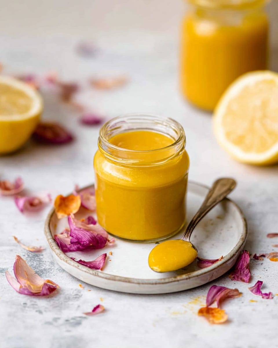 A small clear glass jar filled with a thick, smooth, bright yellow sauce sits in the center of a white round plate; next to it, a silver spoon rests on the plate holding a small amount of the same yellow sauce. Around the plate, scattered dry, colorful onion skins in shades of purple, pink, and orange add texture and color contrast. The background features a white marbled texture surface, with a lemon cut in half on the right side and another glass jar filled with the same yellow sauce slightly blurred in the background. photo taken with an iphone --ar 4:5 --v 7