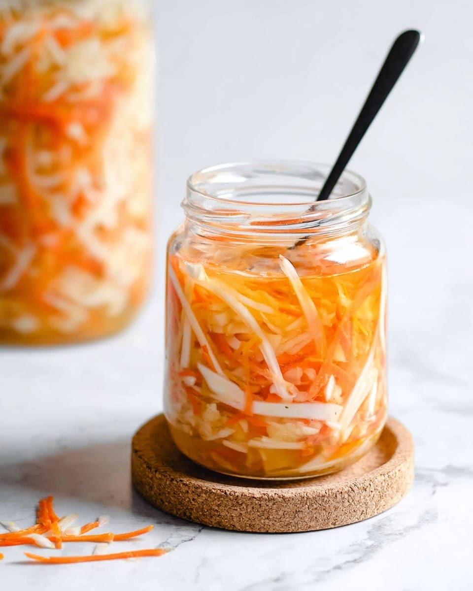 A clear glass jar filled with thin, shredded strips of white and orange vegetables, soaked in a light orange liquid, showing a mix of crunchy textures inside. The jar sits on a round cork coaster placed on a white marbled surface, with some extra shredded vegetable pieces scattered nearby. A black straw is placed inside the jar, leading out of the frame. In the background, another similar jar filled with the same mix is slightly blurred. photo taken with an iphone --ar 4:5 --v 7