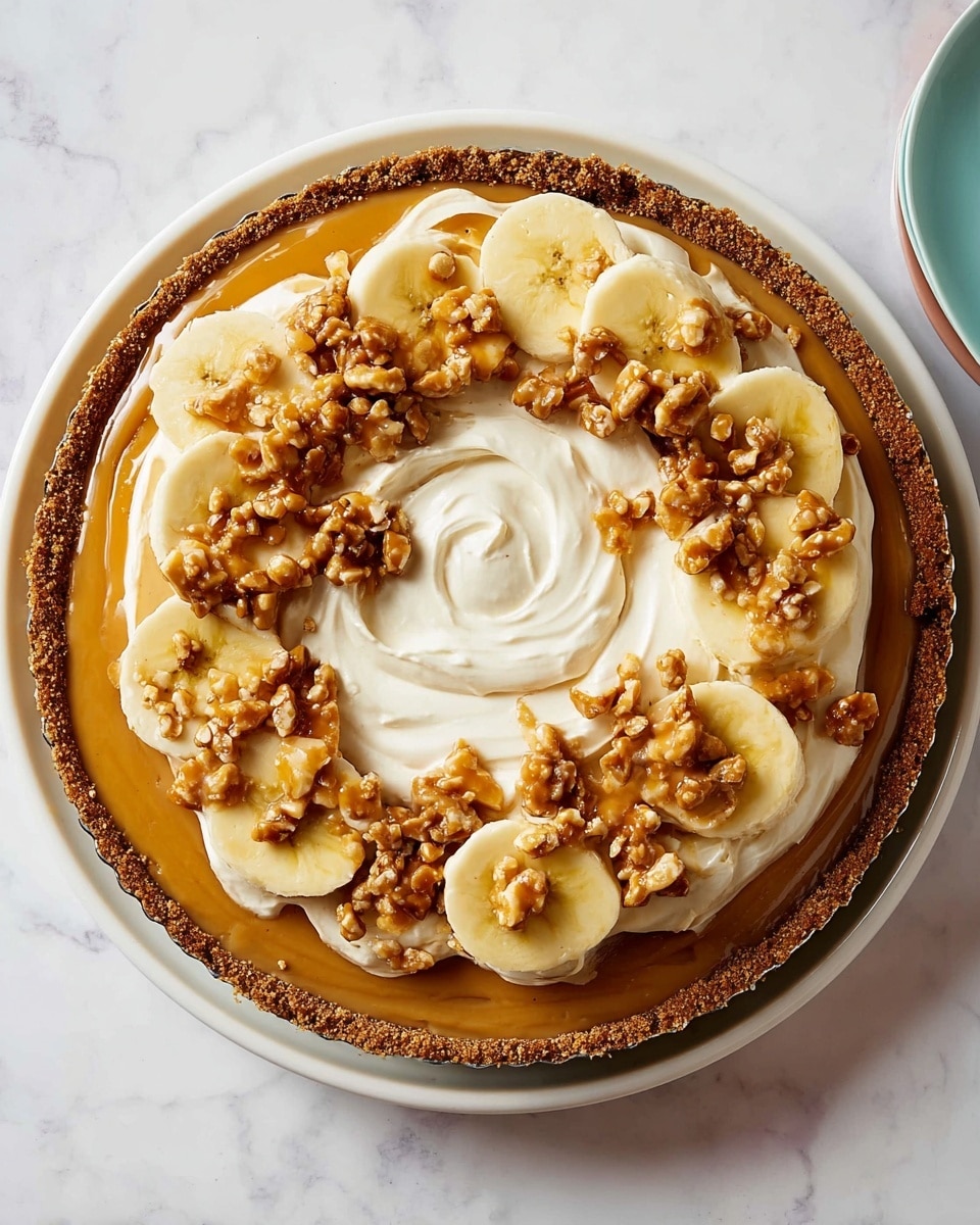 This dessert has four main layers starting from the bottom with a thick, crumbly brown crust that holds the pie together. Above the crust is a smooth, caramel-colored layer spreading evenly across the base. Next, there is a visible ring of fresh banana slices placed around the edge. On top of the bananas, a thick, fluffy white cream is spread in a swirling pattern covering the whole surface. Finally, small clusters of crunchy caramel-coated nuts are scattered over the cream for a textured finish. The dessert is served on a white plate, all placed on a white marbled background. photo taken with an iphone --ar 4:5 --v 7