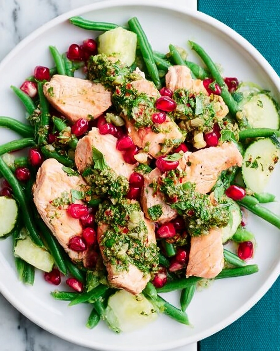 A white plate holds a fresh salad with several layers. The bottom layer is green beans spread across the plate, along with pieces of sliced cucumber on one side. On top of this are medium-sized chunks of cooked salmon with a light orange-pink color. Scattered around the salmon and vegetables are bright red pomegranate seeds adding contrast. The whole dish is topped with a green herb sauce that covers parts of the salmon and some vegetables. The plate rests on a white marbled surface. Photo taken with an iphone --ar 4:5 --v 7