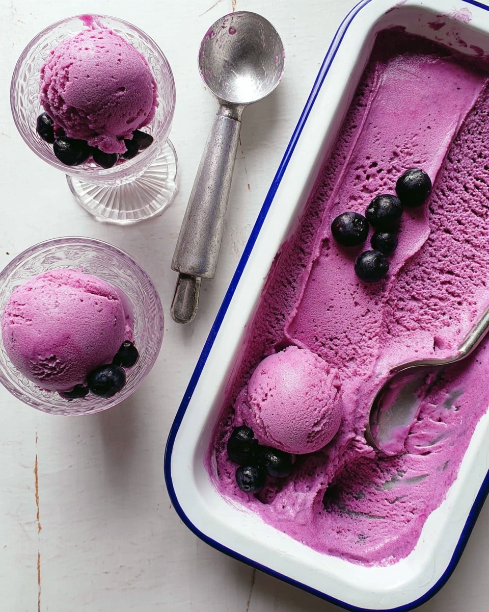 The image shows a white enamel tray with a blue rim filled with bright purple ice cream that looks smooth and creamy with some texture from small ice crystals. There are a few black blueberries placed on top of the ice cream in the tray. Next to the tray, there is a silver ice cream scoop with some purple ice cream left in it. Two clear glass cups filled with one scoop each of the same bright purple ice cream are placed beside the scoop, each topped with a few black blueberries. The background surface is a white marbled texture. photo taken with an iphone --ar 4:5 --v 7