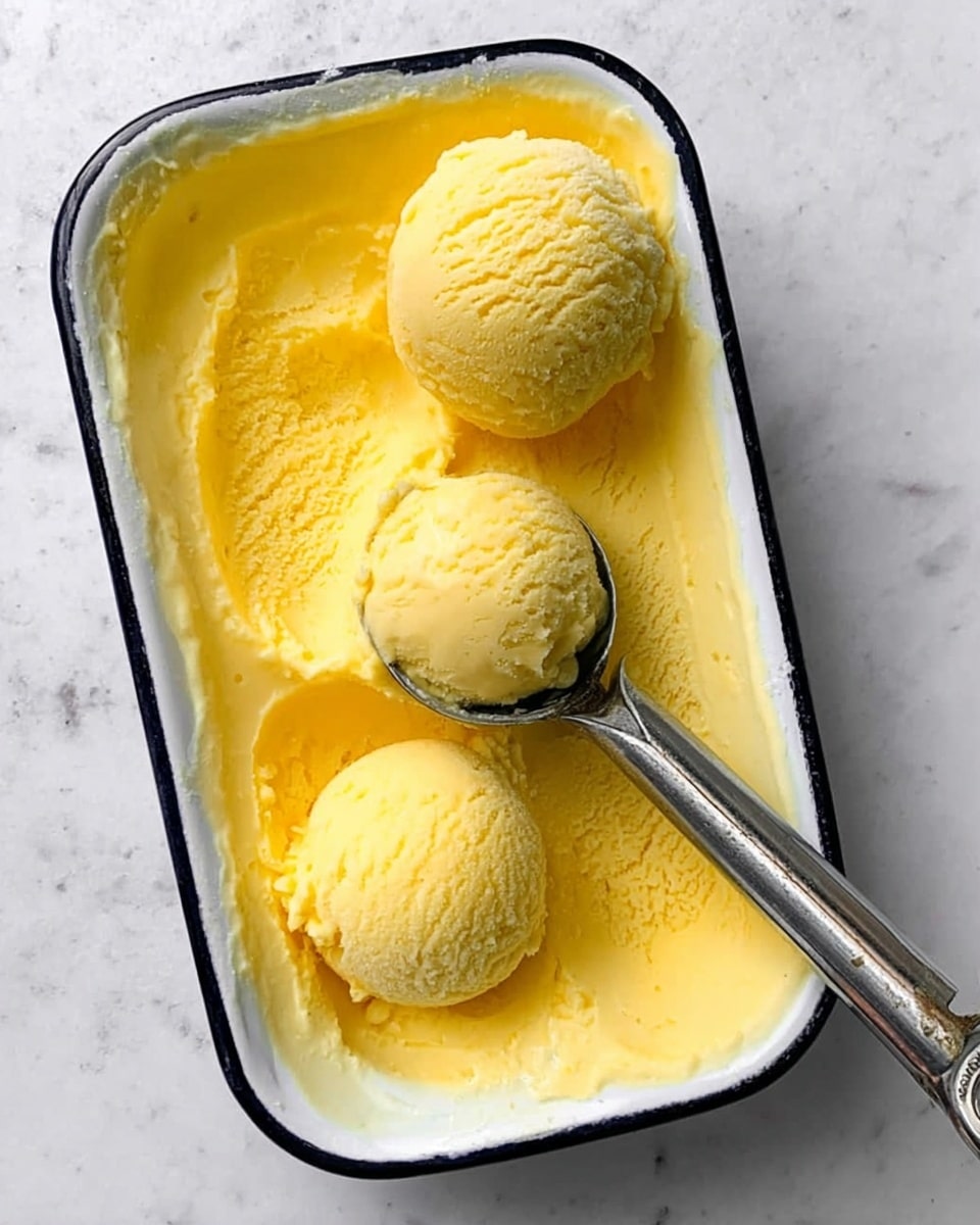 The image shows a white rectangular dish with a black rim, filled with bright yellow ice cream that has a smooth, creamy texture. Within the dish, there are three round scoops of the ice cream, two scoops resting on the surface and one scoop held by a silver ice cream scooper positioned near the bottom right. The white marbled surface background adds a clean and fresh look to the scene. photo taken with an iphone --ar 4:5 --v 7