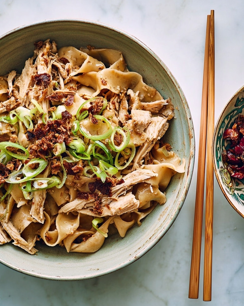 A bowl of wide, flat noodles in a soft light brown color lies at the base, mixed with shredded light beige chicken pieces. Crispy, dark brown bits are scattered over the top, adding texture, while thin, bright green slices of scallion sit atop, providing a fresh contrast. Two light wooden chopsticks rest across the rim of the bowl with a white marbled background setting a clean scene. Photo taken with an iphone --ar 4:5 --v 7