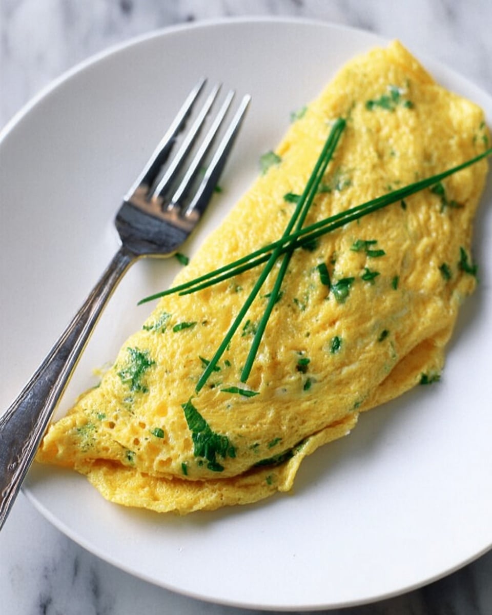 The image shows a yellow omelette folded neatly on a white plate with a smooth and slightly glossy texture, speckled with green herbs inside, and garnished on top with three long green chives arranged in an X shape. A silver fork lies next to the omelette on the plate, with the plate placed on a white marbled surface. Photo taken with an iphone --ar 4:5 --v 7