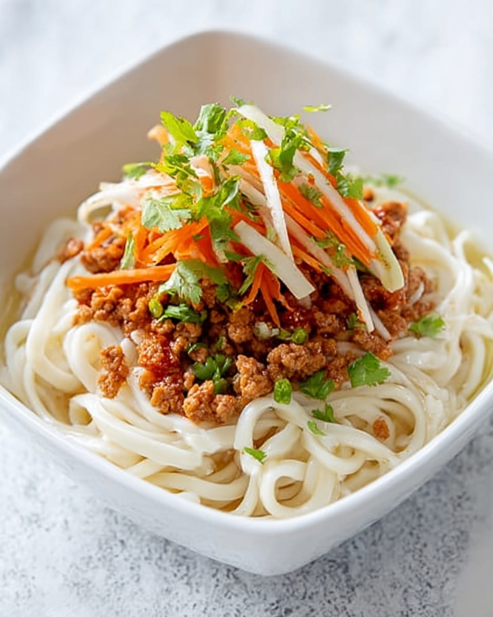 A square white bowl holds a serving of thick, white noodles at the bottom, topped with small chunks of browned minced meat spread evenly across the noodles. On top of the meat is a colorful pile of thinly sliced vegetables, including orange carrot strips and pale white slices, both with a fresh green herb garnish scattered over them. The dish sits on a white marbled surface with soft natural lighting highlighting the textures and colors. Photo taken with an iphone --ar 4:5 --v 7