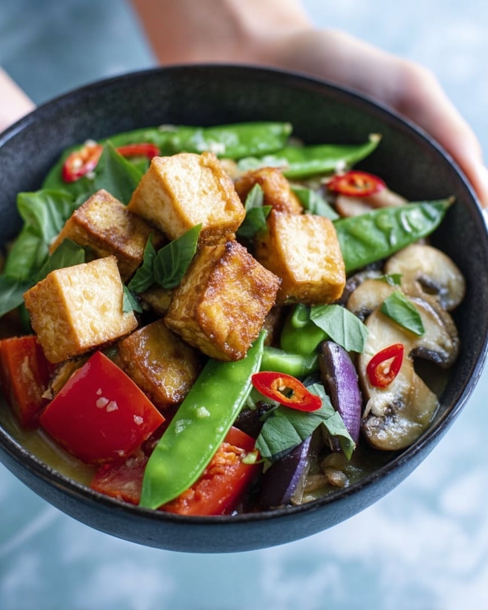 A close-up of a black bowl held by a woman's hand, filled with a colorful mix of food. There are multiple layers starting with green snap peas and fresh green basil leaves scattered on top. Next, the dish shows light brown fried tofu cubes with a crispy texture standing upright. Around these are soft mushroom slices with a light beige color, along with chopped red chili rings adding a bright red contrast. There are also chunks of light purple eggplant and red bell pepper pieces. The bowl sits against a soft blue background, and the woman's hand is gently holding it from the bottom. The surface beneath is a white marbled texture. photo taken with an iphone --ar 4:5 --v 7