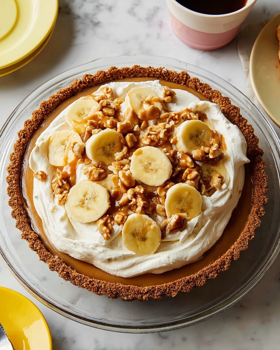A round tart with a thick, crumbly brown crust holds several layers. The bottom layer is smooth caramel spread evenly to the edge of the crust. On top of the caramel, there are several slices of yellow banana arranged close to the crust, visible around the edge. Next, a thick, fluffy layer of white whipped cream covers the bananas and caramel in swirls. Scattered on top of the whipped cream are small clusters of caramel-coated nuts, some whole and some broken into pieces, adding a crunchy texture. The tart is placed on a transparent plate on a white marbled surface, with partial views of a cup of coffee and two yellow plates nearby. Photo taken with an iphone --ar 4:5 --v 7