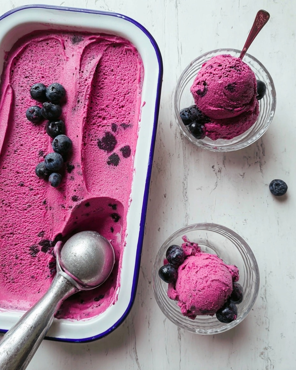 The image shows a large white dish with a thin dark blue edge holding a bright magenta ice cream spread flat, with some fresh dark blueberries placed on top near the edges. Next to the dish lies a metal ice cream scoop with some of the magenta ice cream still inside the scoop’s bowl. To the right, there are two clear glass bowls with one scoop each of the same magenta ice cream, each scoop topped with a few dark blueberries. The background is a white marbled surface. photo taken with an iphone --ar 4:5 --v 7