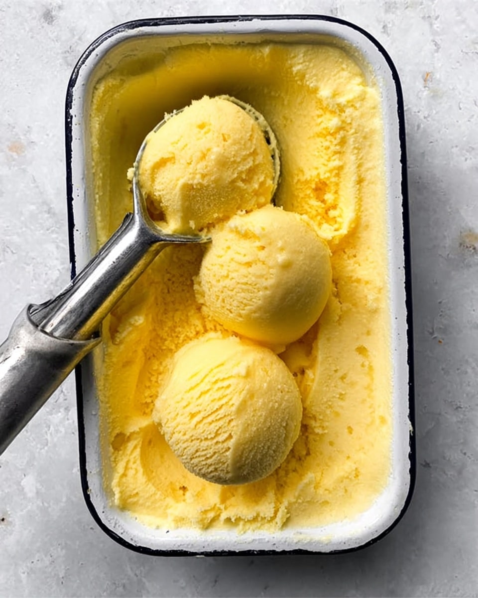 The image shows a white metal tray with a black rim placed on a white marbled surface. Inside the tray, there is a smooth, creamy yellow ice cream or sorbet base. Three round scoops of the same yellow frozen dessert rest on top, with one being held slightly above the tray by a silver ice cream scooper. The texture of the dessert appears soft and slightly fluffy, with some melting around the edges. photo taken with an iphone --ar 4:5 --v 7