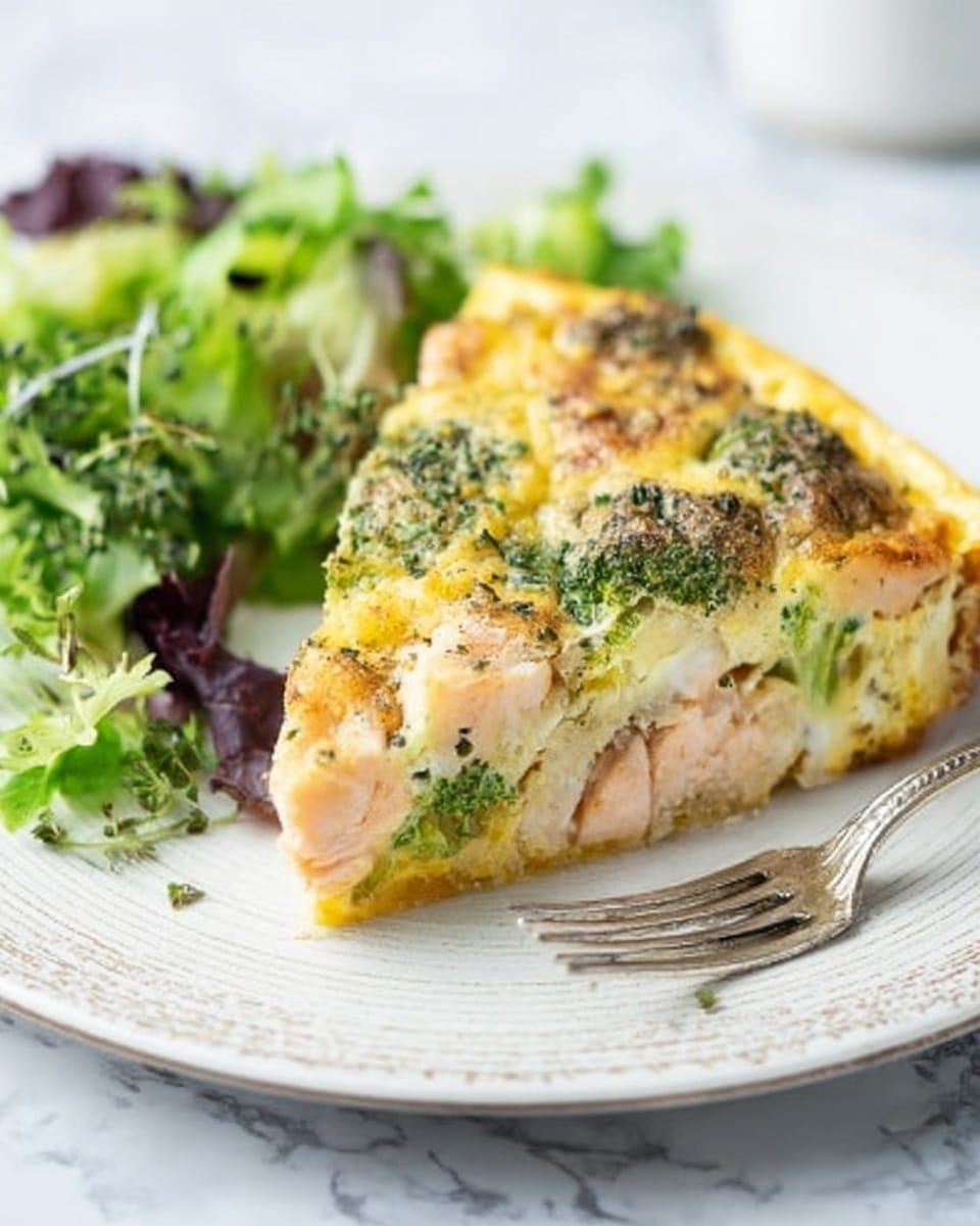 A slice of frittata sits on a white plate with a delicate fork beside it. The frittata is thick with several layers: a golden-yellow egg base, chunks of light pink cooked salmon, green broccoli florets dotted across the top, and a sprinkling of herbs giving a textured, slightly crispy look on top. To the side of the frittata slice is a small pile of fresh mixed greens with light and dark green leaves. The plate rests on a white marbled surface, creating a clean and bright setting. photo taken with an iphone --ar 4:5 --v 7