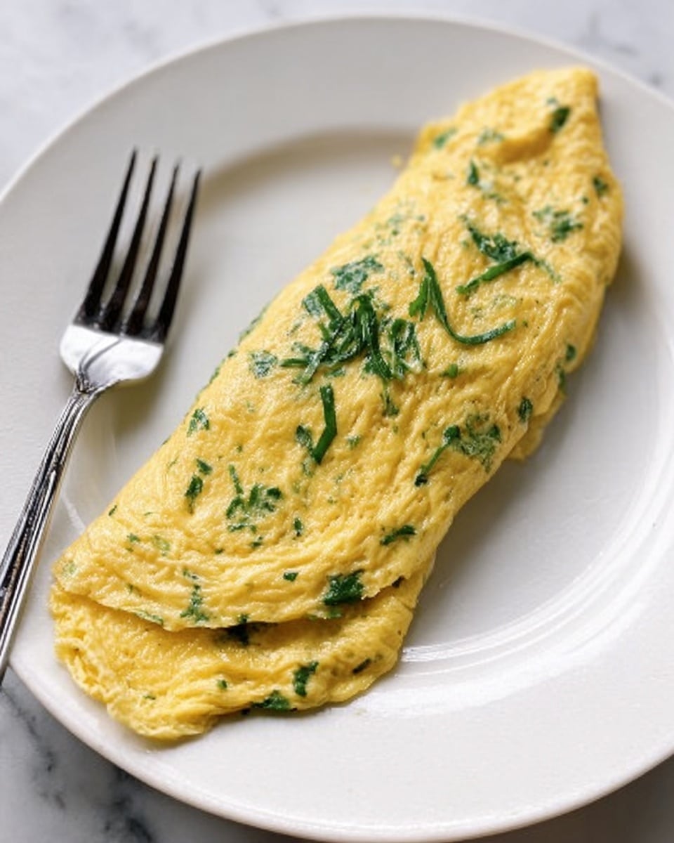A smooth, folded yellow omelette with green herbs mixed inside sits centered on a clean white plate. The omelette has a soft, slightly glossy texture with visible pieces of herbs throughout. A silver fork rests diagonally in front of the omelette on the plate. The background is a white marbled surface. photo taken with an iphone --ar 4:5 --v 7