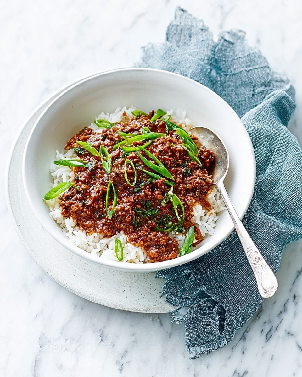 A white bowl filled with a layer of white rice at the bottom, topped with a thick, textured brown meat sauce scattered unevenly over it. The sauce has small visible bits of meat and is sprinkled with thin green slices of spring onions or chives, adding a fresh pop of color. The bowl sits on a larger white plate with a silver spoon inside, resting on the bowl’s edge, all placed on a white marbled surface. Next to the bowl is a crumpled blue-grey cloth napkin. Photo taken with an iphone --ar 4:5 --v 7