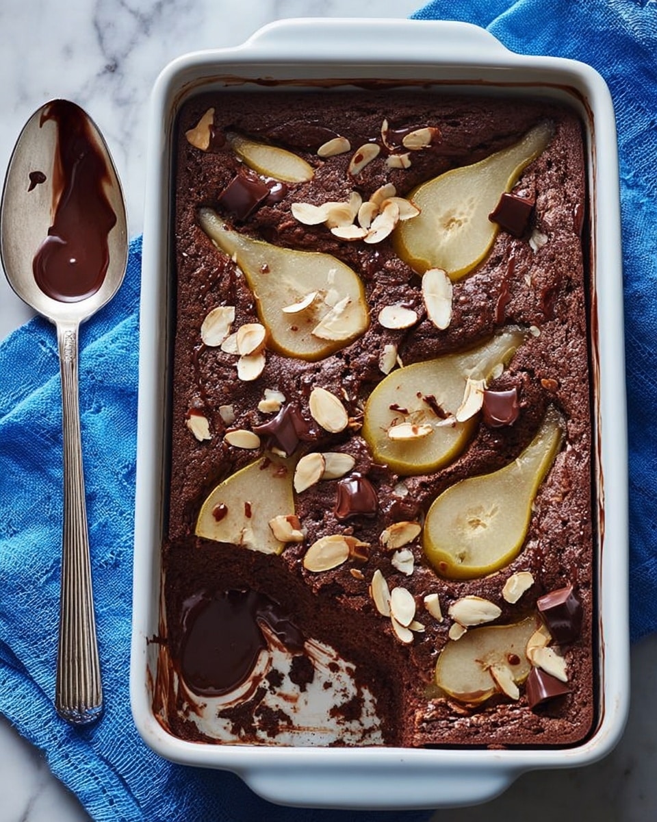 A white rectangular baking dish holds one layer of moist, dark brown chocolate cake with a rough texture, embedded with sliced pale yellow pears and scattered with toasted light tan almond flakes. Large chunks of melted dark chocolate are also spread throughout the cake, some sunken into the surface. A silver spoon with chocolate residue rests above the dish on a white marbled texture. Part of the cake is scooped out from the top left corner, revealing gooey chocolate beneath. photo taken with an iphone --ar 4:5 --v 7