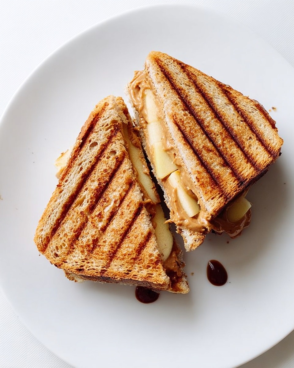 A grilled sandwich cut diagonally into two halves is placed on a white plate. The sandwich has two slices of golden-brown toasted bread with grill marks running vertically. Inside, the top visible layer shows melted cheese with a smooth, creamy texture, followed by a darker layer of thinly sliced apples and a spread of peanut butter oozing slightly. The plate sits on a white marbled surface. photo taken with an iphone --ar 4:5 --v 7