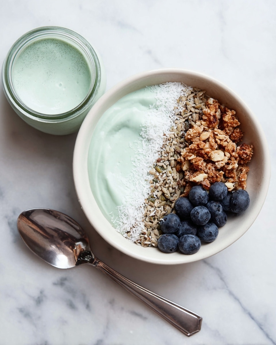 A white bowl holds a layered dish with four distinct parts: on the left, a smooth, pale green creamy base; next to it, a thin line of white shredded coconut; above that, a layer of mixed granola with seeds and nuts showing a rough texture; and on the bottom right, a cluster of fresh, plump dark blue blueberries. The bowl is placed on a white marbled texture surface with a spoon lying nearby and a small glass jar filled with the same pale green cream to the right. Photo taken with an iphone --ar 4:5 --v 7