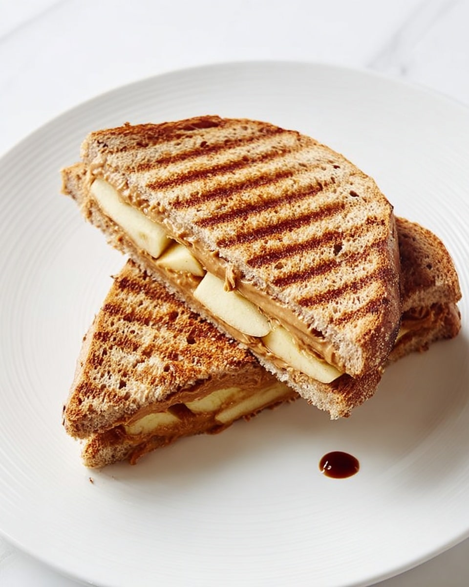 A grilled sandwich cut diagonally into two halves, placed on a white plate with a white marbled texture beneath. The sandwich has three main layers: the outer layers are toasted brown bread with clear grill marks and a slightly rough texture, the middle layer contains smooth peanut butter with a light brown color, and slices of pale yellow apple with a slight shine, some pieces peeking out from the edge of the bread. The sandwich halves are positioned so their cut sides face upward, showing the filling clearly. A small dark drop sits on the plate near the sandwich. Photo taken with an iphone --ar 4:5 --v 7