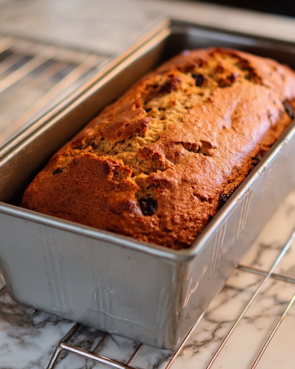 The image shows a freshly baked loaf of bread inside a silver metal bread pan on a white marbled surface. The bread has a golden brown crust with darker spots where ingredients like nuts or dried fruits are visible. The top of the bread is slightly cracked and has a rough texture, showing it just came out of the oven. The bread pan is placed on a metal rack, which is part of the oven setup. Photo taken with an iphone --ar 4:5 --v 7