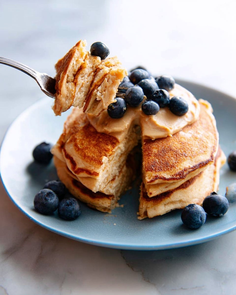 A stack of three golden-brown pancakes sits on a white plate with a slightly rough texture. The pancakes have a fluffy, slightly uneven surface showing their soft texture. On top of the stack is a thick layer of light brown peanut butter, partly melted, with several fresh, plump blueberries scattered both on the peanut butter and around the plate. A silver fork, holding three small triangular pancake pieces, is near the stack, with one blueberry beside the fork. The background is a white marbled texture. photo taken with an iphone --ar 4:5 --v 7