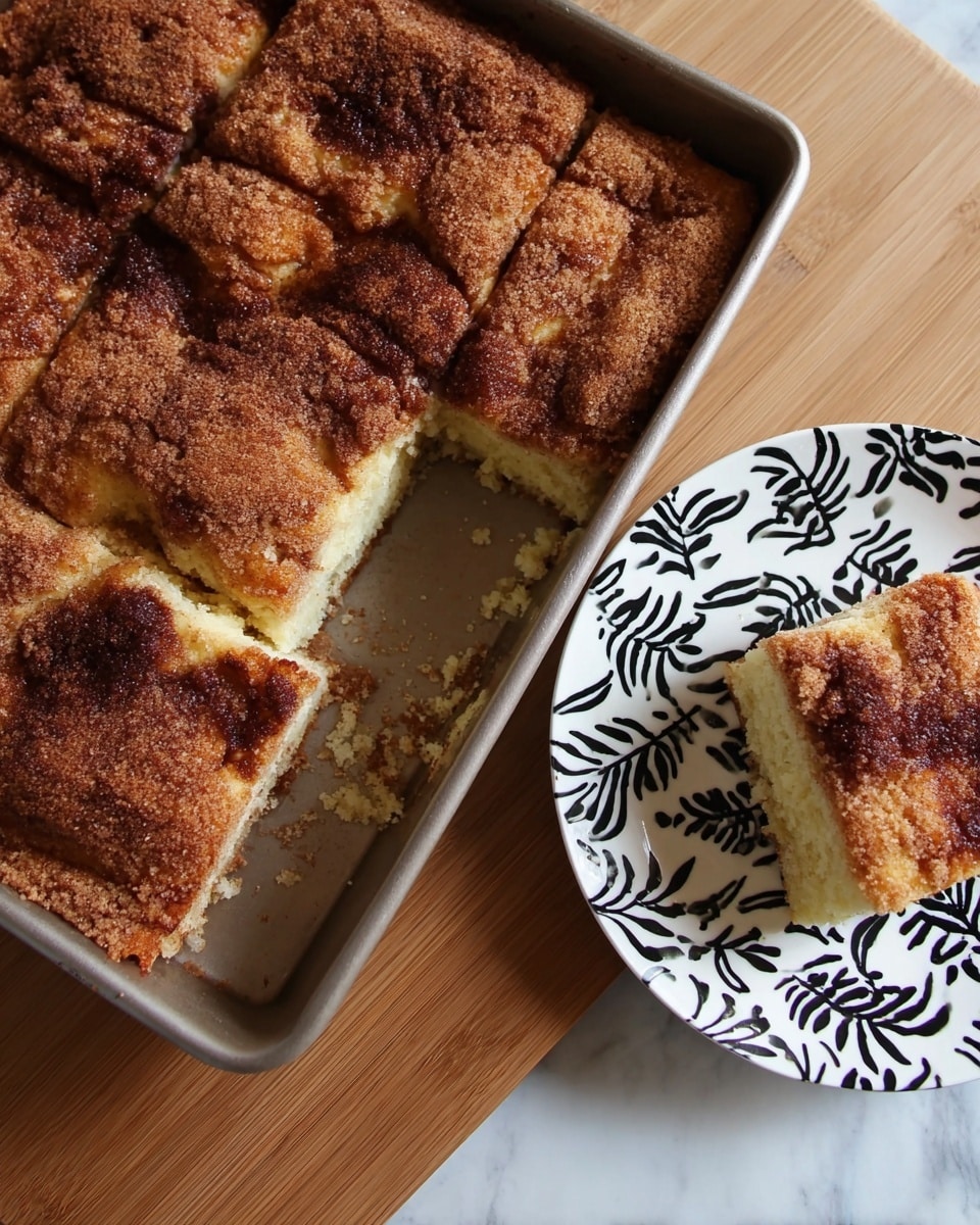 The image shows a thick, square piece of coffee cake with a golden-brown crust and a crumbly, cinnamon-sugar topping. The cake itself is light yellow in color with a soft and fluffy texture visible on the cut sides. The crumb topping is unevenly spread, with some darker brown spots showing the cinnamon mixed in. The cake is placed on a white plate with a black floral and leaf pattern, which rests on a wooden surface. Next to the plate is a metal baking pan with several more square pieces of the same cake, with one piece missing from the corner, revealing the soft inside. photo taken with an iphone --ar 4:5 --v 7