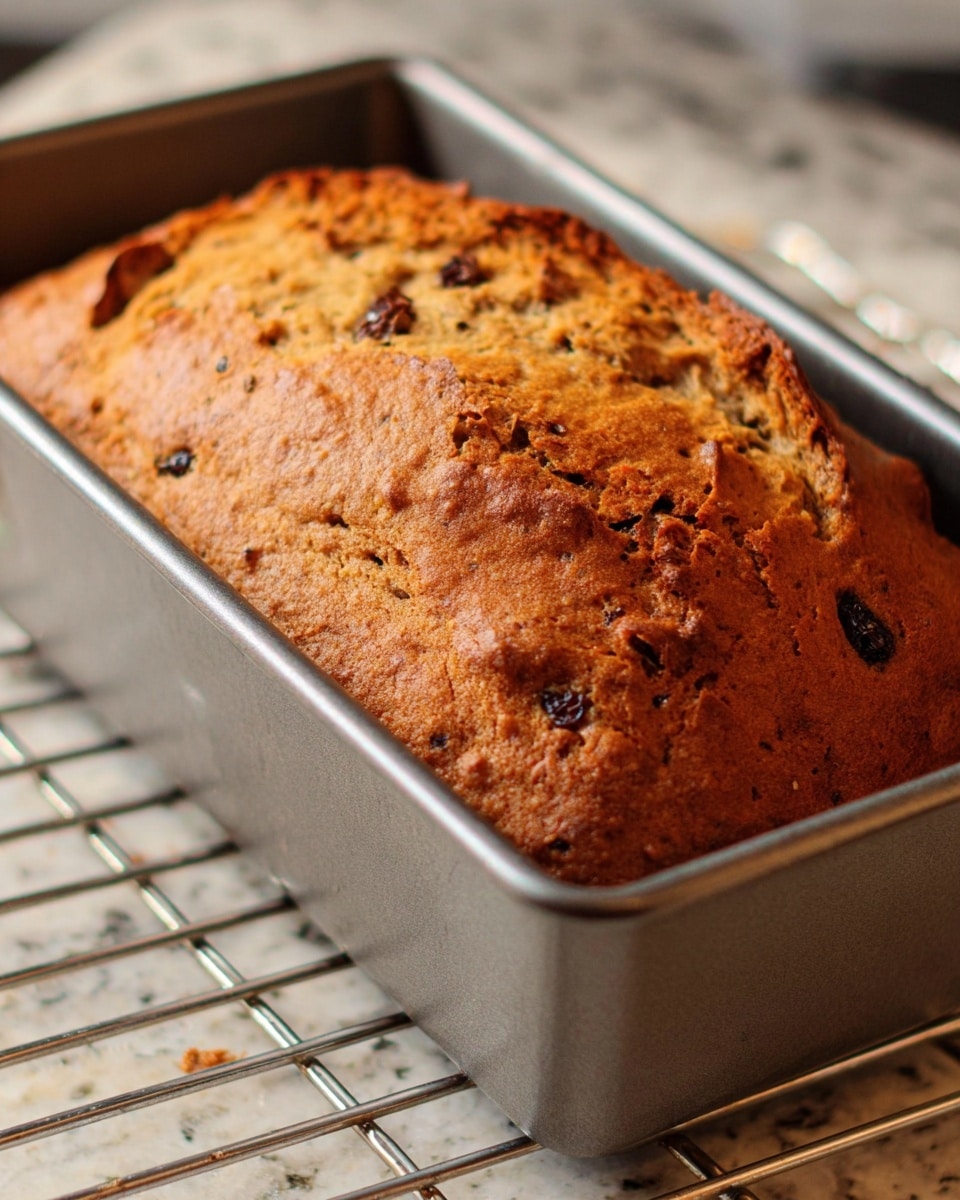 The image shows a freshly baked bread loaf in a silver metal baking pan, placed on a wire rack inside an oven. The bread has a golden-brown crust with small dark spots, possibly from chocolate chips or raisins, and a slightly cracked surface revealing a soft inside. The metal pan has straight vertical ridges on its sides. The background is a white marbled texture. Photo taken with an iphone --ar 4:5 --v 7