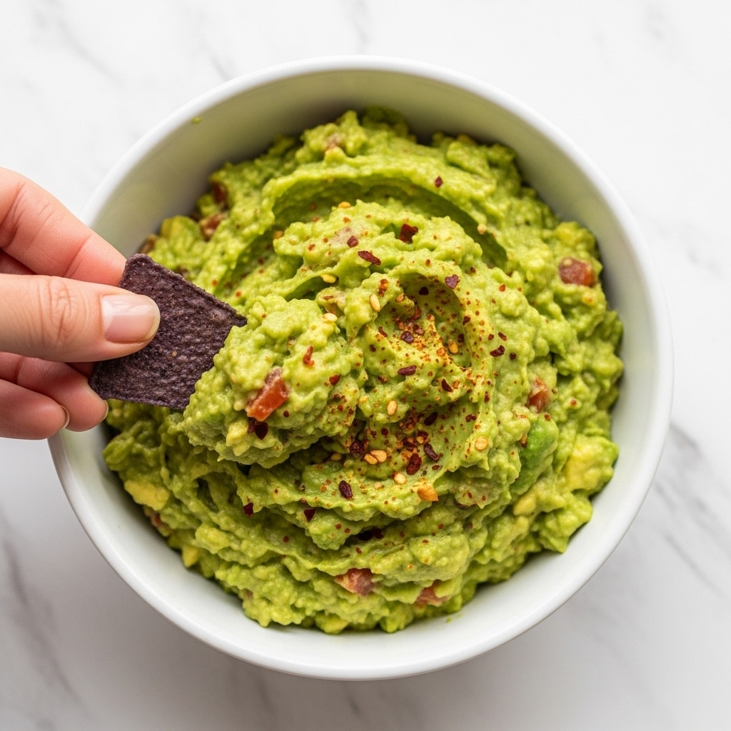 A white bowl filled with chunky green guacamole, showing bits of avocado and small red seasoning flakes on top. A woman's hand is holding a blue corn tortilla chip just above the guacamole, ready to dip. The background is a white marbled surface. Photo taken with an iphone --ar 4:5 --v 7