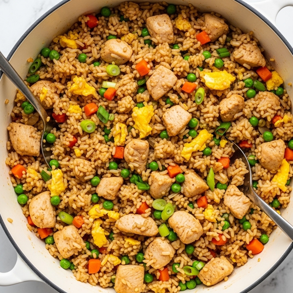 The image shows a close-up view of a pot filled with fried rice mixed evenly with small pieces of cooked chicken, scrambled egg bits, green peas, diced orange carrots, and chopped green onions. The fried rice is golden brown with a slightly glossy texture, and the chicken pieces are light brown and tender-looking. A metal spoon and tongs rest inside the pot against the food. The pot is on a white marbled surface. photo taken with an iphone --ar 4:5 --v 7