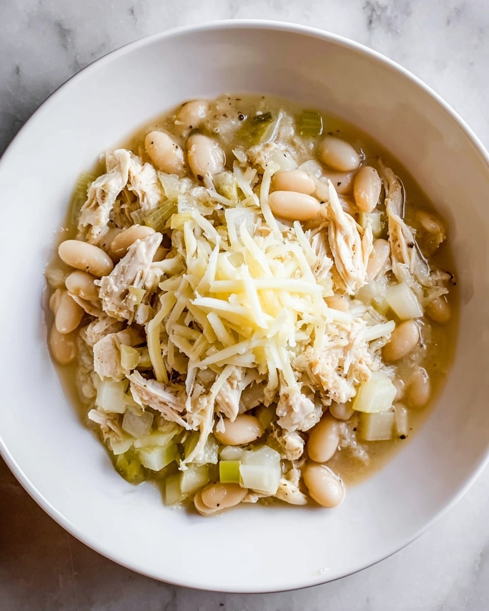 The dish is served in a white bowl filled with several layers. The bottom layer shows a light, slightly thick sauce with a pale green tint, mixed with chunks of shredded white chicken and small white beans scattered throughout. Mixed in are small greenish cubes, possibly vegetables, adding texture and color contrast. On top of this mixture is a modest pile of shredded white cheese, placed in the center, providing a creamy texture against the savory ingredients beneath. The whole dish rests on a white marbled texture surface. photo taken with an iphone --ar 4:5 --v 7
