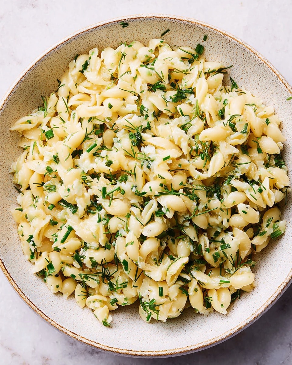 A white bowl filled with a mix of short, pale yellow pasta pieces that have a soft texture, scattered evenly throughout the dish. Small green herb leaves and finely chopped green chives are mixed in, adding contrast and freshness. The bowl is placed on a white marbled surface, and the overall look is bright and simple without extra layers. photo taken with an iphone --ar 4:5 --v 7