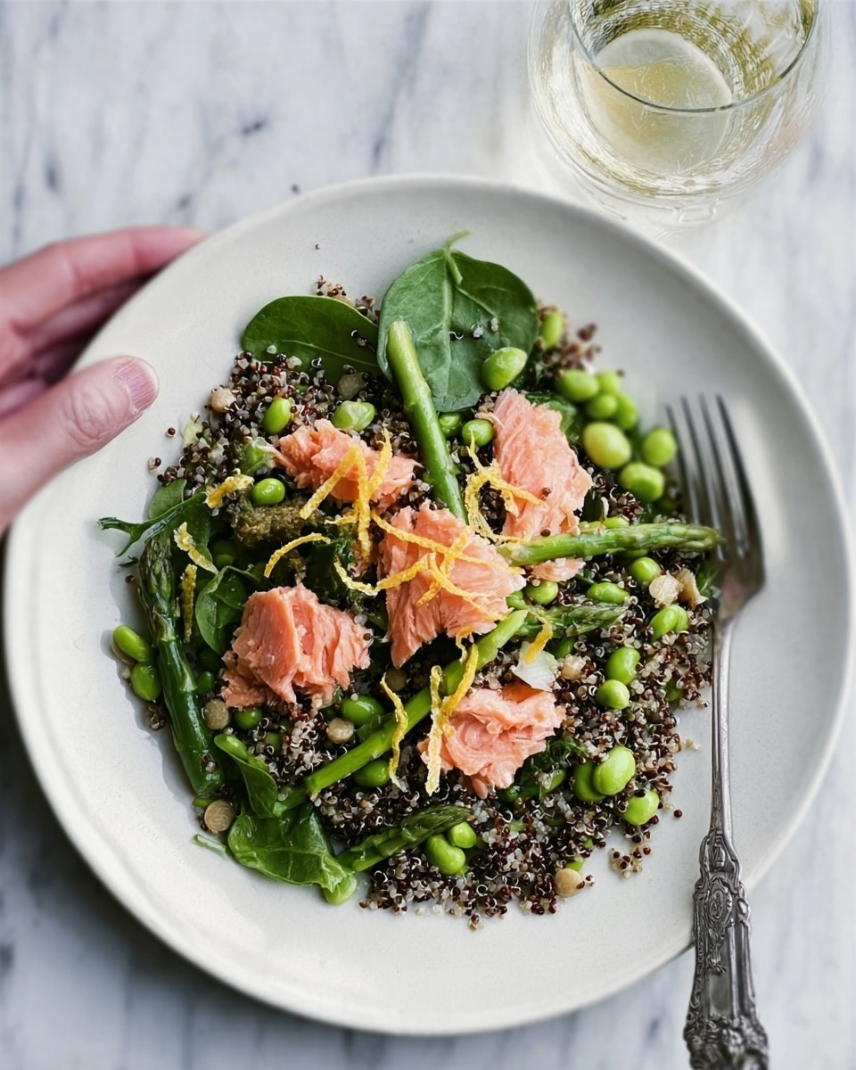 A white plate holds a fresh salad arranged in layers. The bottom layer is a mix of dark green spinach leaves scattered around. On top of that, there is a layer of black quinoa mixed with green edamame beans, creating a rough, grainy texture. Bright green asparagus spears lie across one side of the plate, slightly overlapping the quinoa. Pieces of pink cooked salmon are placed in small clusters over the greens and grains. Thin yellow lemon zest strips are delicately placed on the salmon. The salad is garnished with tiny seeds and herbs, adding small brown and green flecks. The plate is set on a white marbled surface with a silver fork beside it and part of a clear glass visible at the top right corner. Photo taken with an iphone --ar 4:5 --v 7