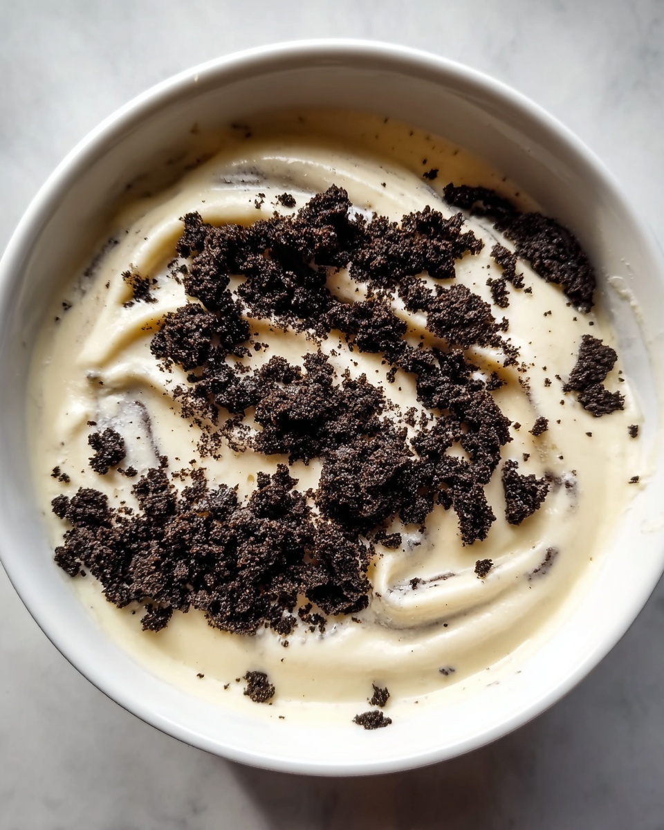 The image shows a white bowl filled with a creamy, smooth layer of light beige pudding or yogurt. On top of this creamy base, there is a generous sprinkle of dark brown, crumbly cookie pieces unevenly spread, creating a rough texture contrast against the smooth cream below. The cookie crumbs vary in size, from fine dust to larger chunks, and are primarily concentrated in the center with some scattered around. The bowl sits on a surface with a white marbled texture, enhancing the dish's clean, simple look. photo taken with an iphone --ar 4:5 --v 7