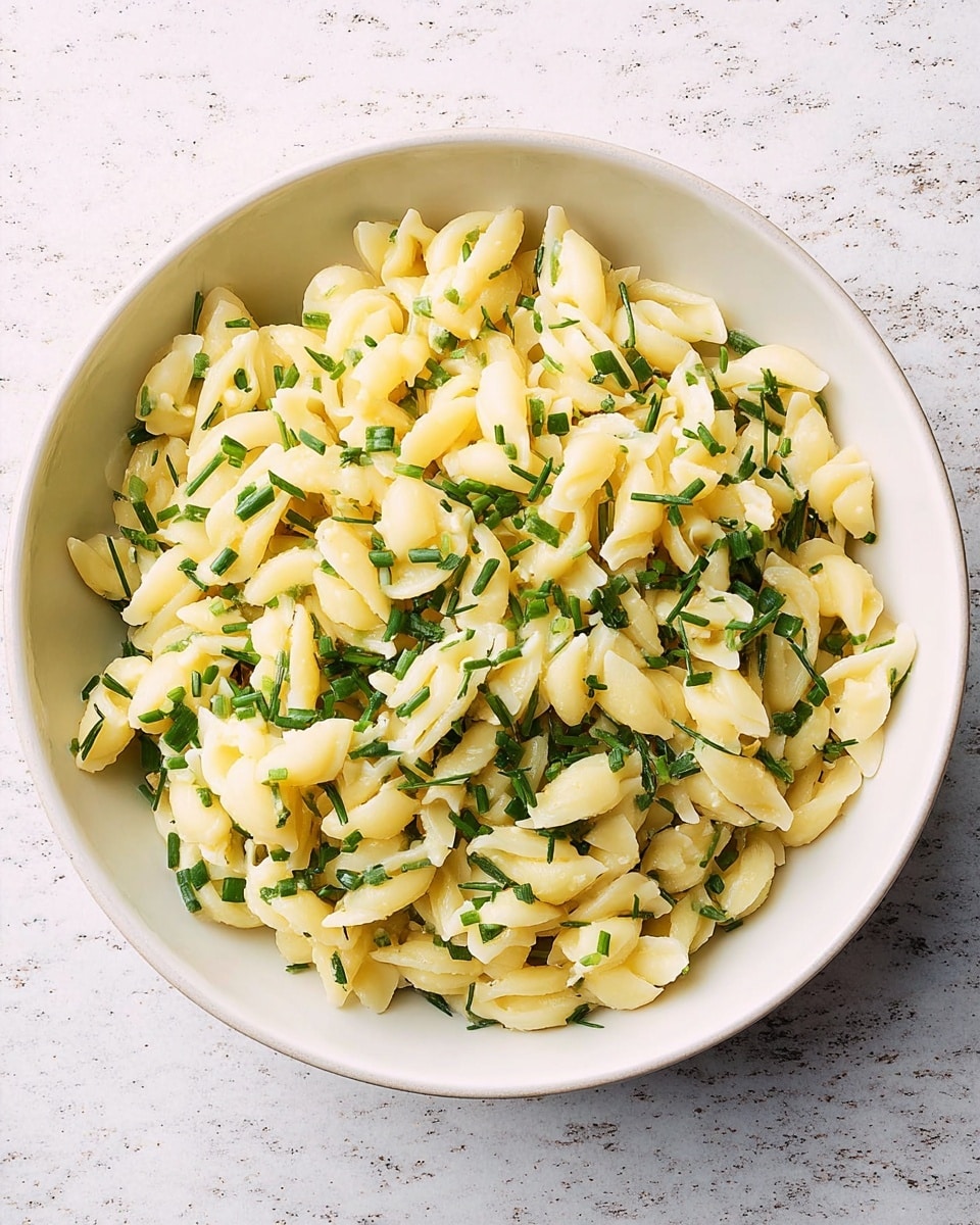 A close-up view of a bowl filled with small, short pasta pieces that are pale yellow in color, mixed with finely chopped green herbs like dill and chives scattered all over. The bowl is white and round with a slightly speckled texture and a darker rim. The pasta’s texture looks soft and slightly oily, with herbs evenly spread throughout. The background is a white marbled surface, adding a clean and bright look to the image. photo taken with an iphone --ar 4:5 --v 7