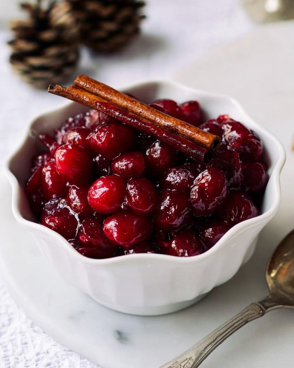 The image shows a white scalloped bowl filled with glossy, deep red cranberries that look cooked or stewed, giving them a soft texture with some juice. On top of the cranberries, there is a single dark brown cinnamon stick placed diagonally. The bowl sits on a white marbled surface with a silver spoon partially visible to the right and some out-of-focus pinecones to the left. The scene has soft lighting, making the colors rich and vibrant. photo taken with an iphone --ar 4:5 --v 7