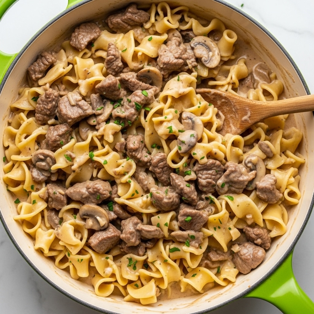 The image shows a close-up of a creamy beef stroganoff dish with thick, flat egg noodles layered in a rich, light brown sauce mixed with tender chunks of browned beef and small pieces of mushrooms. The noodles have a pale yellow color and a soft texture, while the sauce looks smooth and glossy, evenly coating all the ingredients. Small bits of green herbs are sprinkled lightly on top, adding a touch of color contrast to the warm beige tones of the dish. The food is in a white pot with a green handle visible on the side, all set on a white marbled surface. Photo taken with an iphone --ar 4:5 --v 7